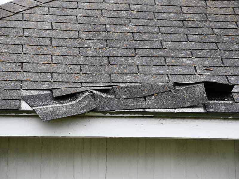 Close up of a residential roof with dark gray shingles that are loose and about to fall off due to storm damage